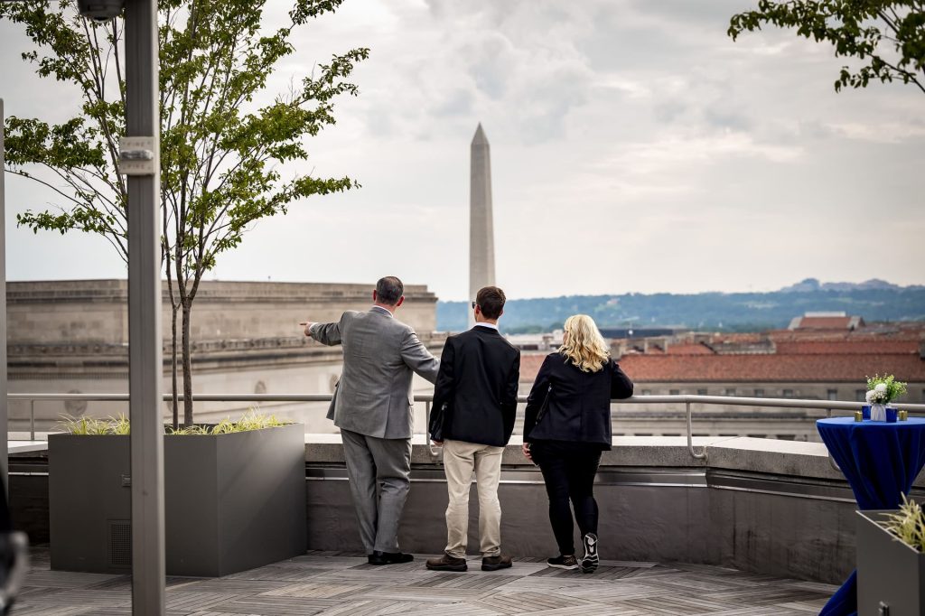Three people on a rooftop terrace looking toward the Washington Monument.