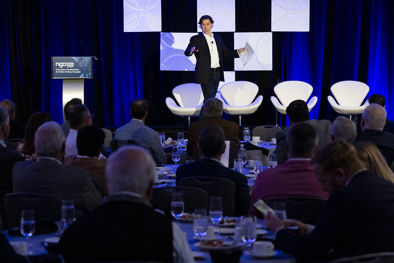 Speaker presenting on stage at an NGA conference, with audience seated at round tables.