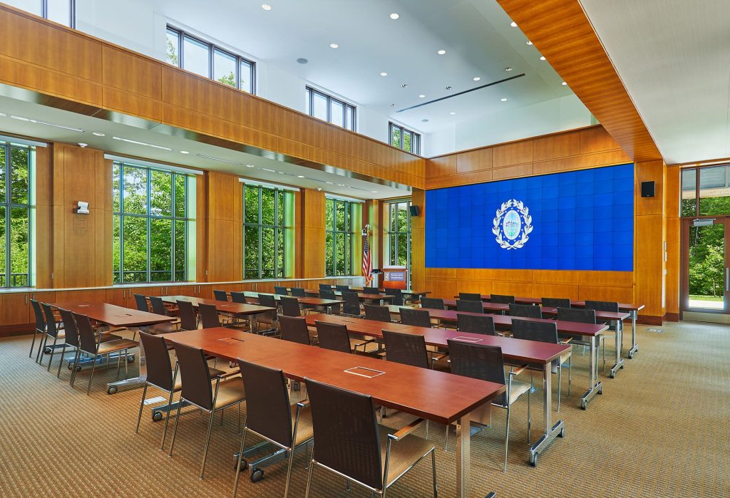 Lecture hall with wood walls, rows of desks, and a large screen at the front.