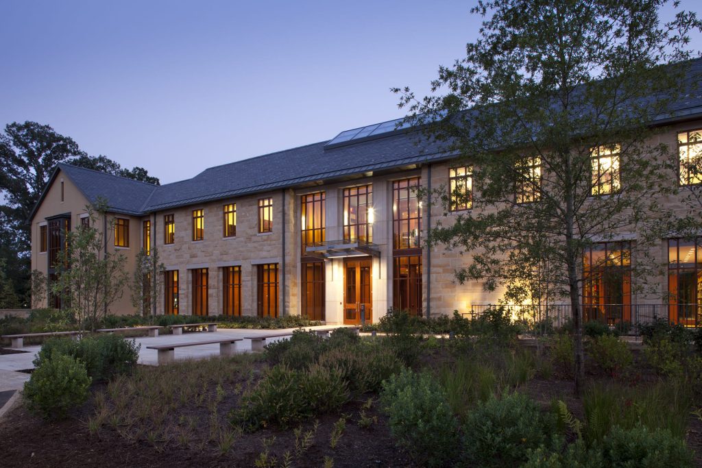 Evening view of a two-story academic building with warm interior lights glowing through tall windows, stone walls, landscaped gardens, and trees in front.