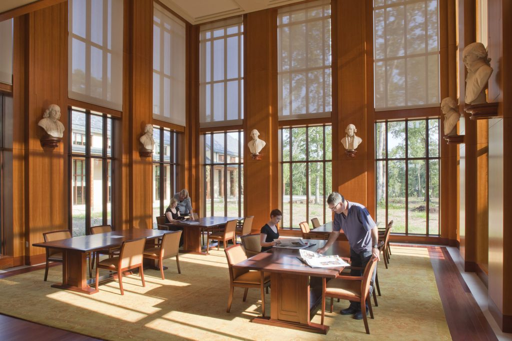 Library or study room with tall wood-paneled walls, large windows letting in natural light, classical busts mounted on the walls, and people reading and working at wooden tables.
